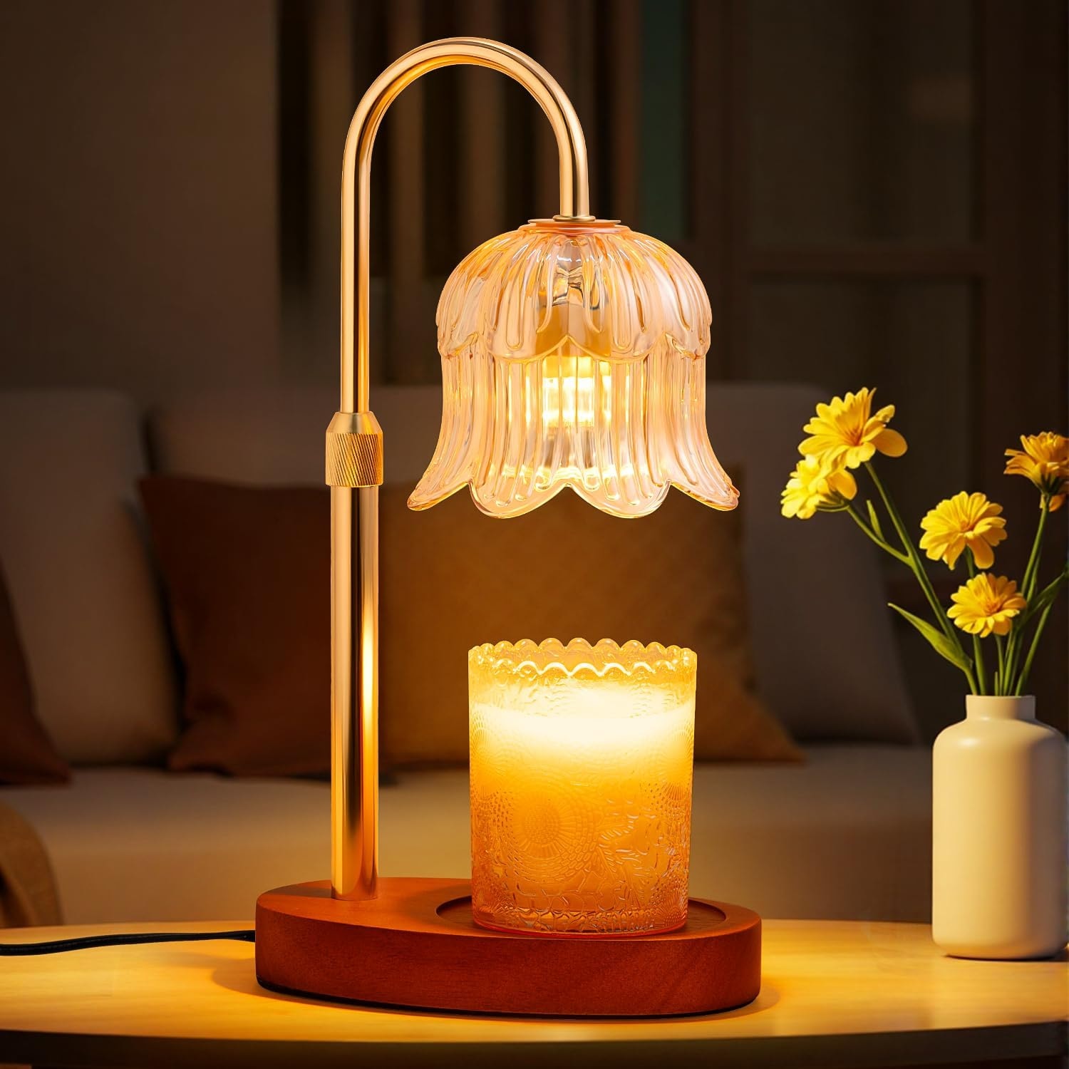 Glass flower candle warmer lamp lit above a jar candle on a wooden table beside yellow flowers