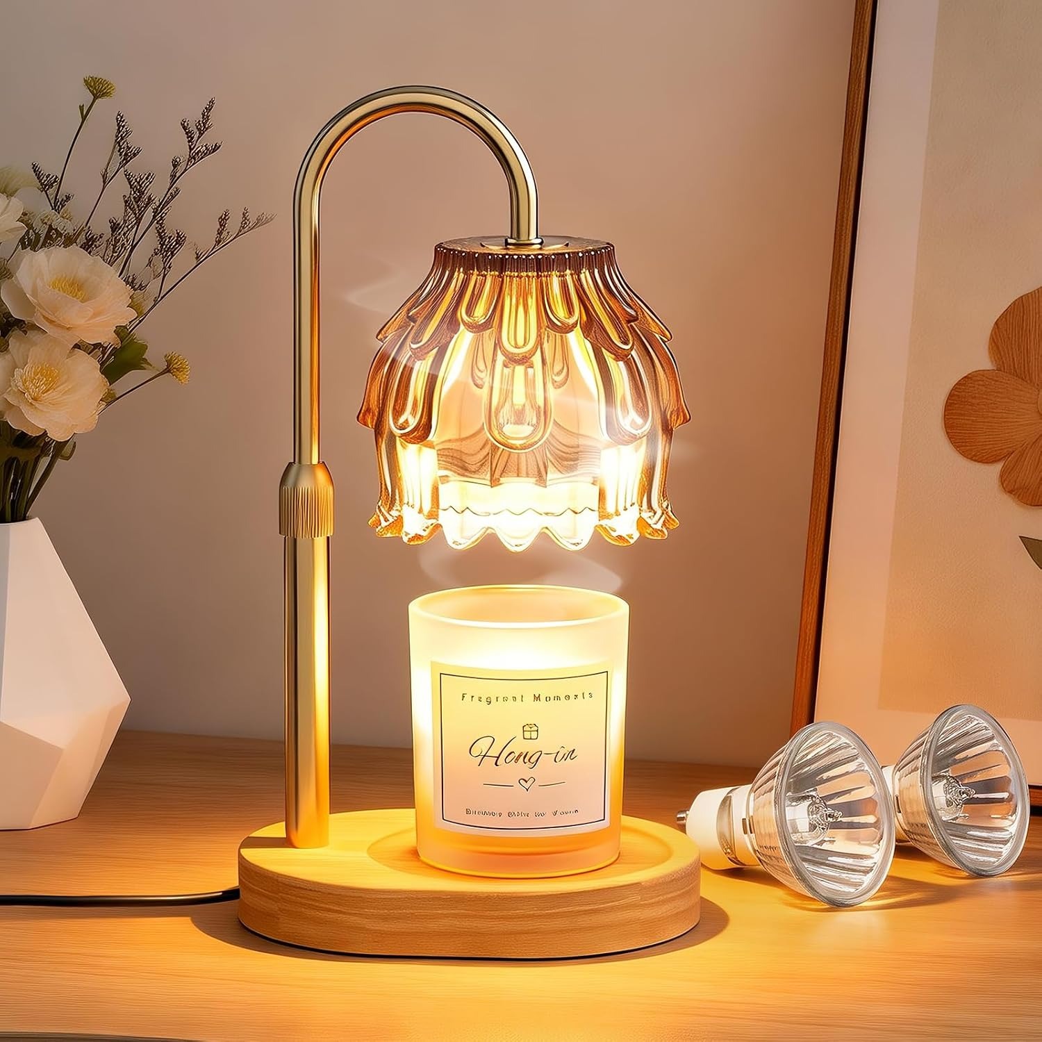 Tawny flower-shaped glass candle warmer lamp with a gold arm and wood base glowing over a white jar candle, shown with two spare GU10 bulbs